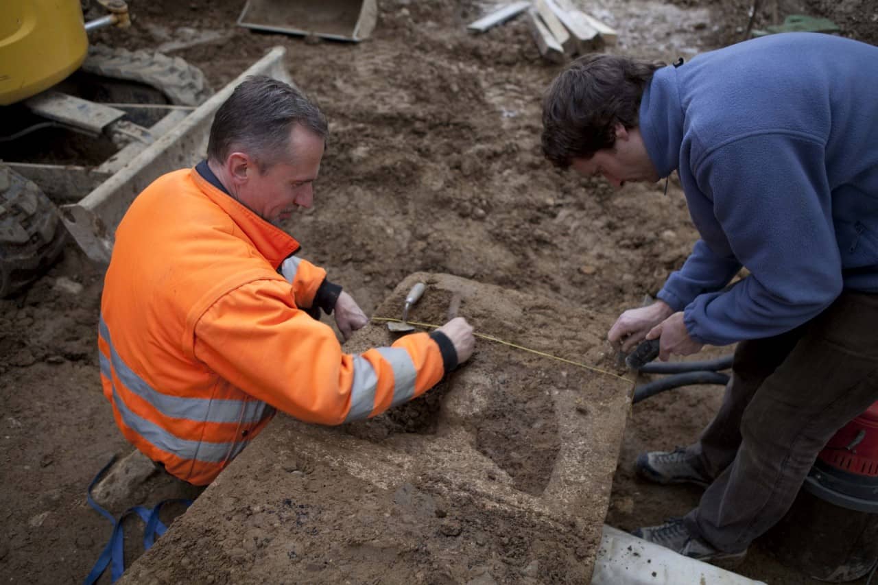 Ein Ausgräber und ein Restaurator säubern den frisch umgedrehten Grabsteins von der Erde.