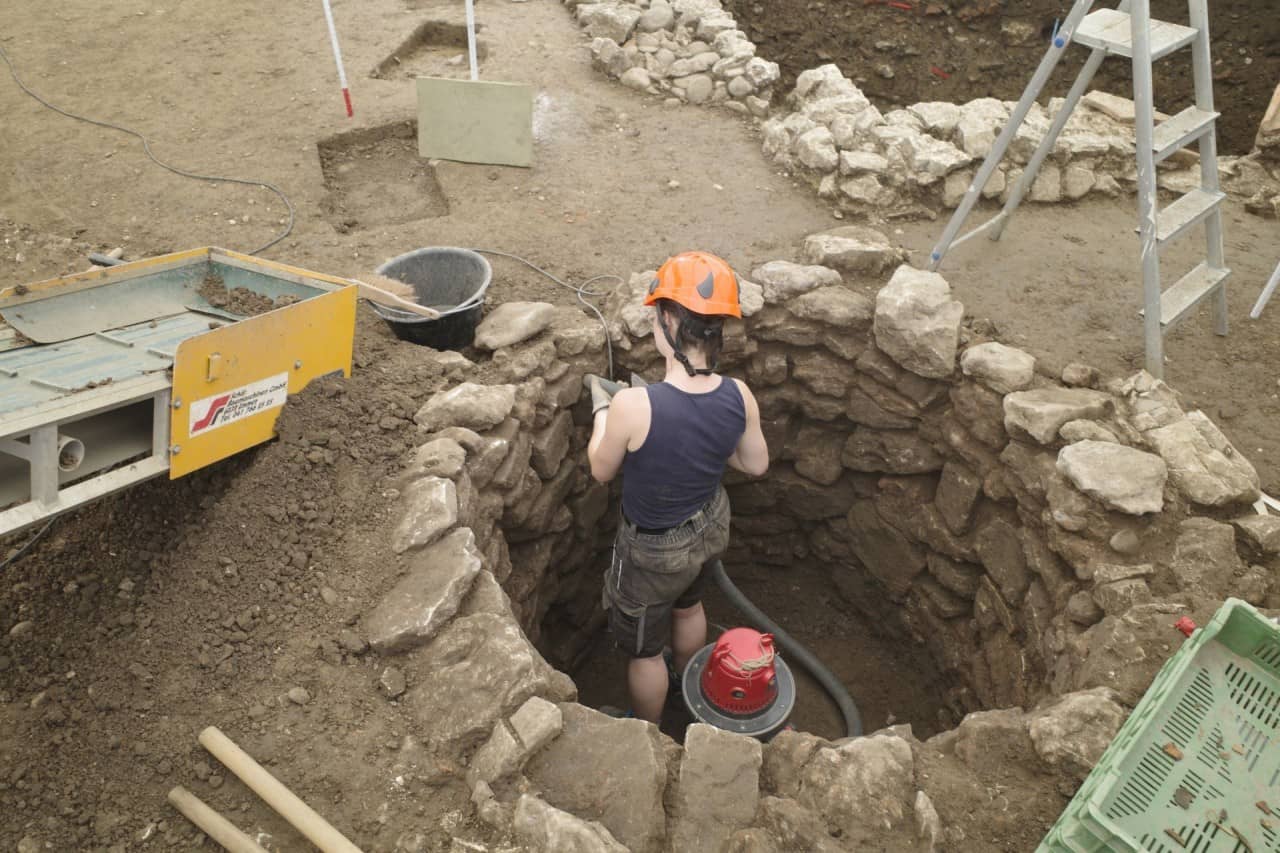 Grabungsmitarbeiterin steht in einem römischen Brunnen und säubert die Mauer.