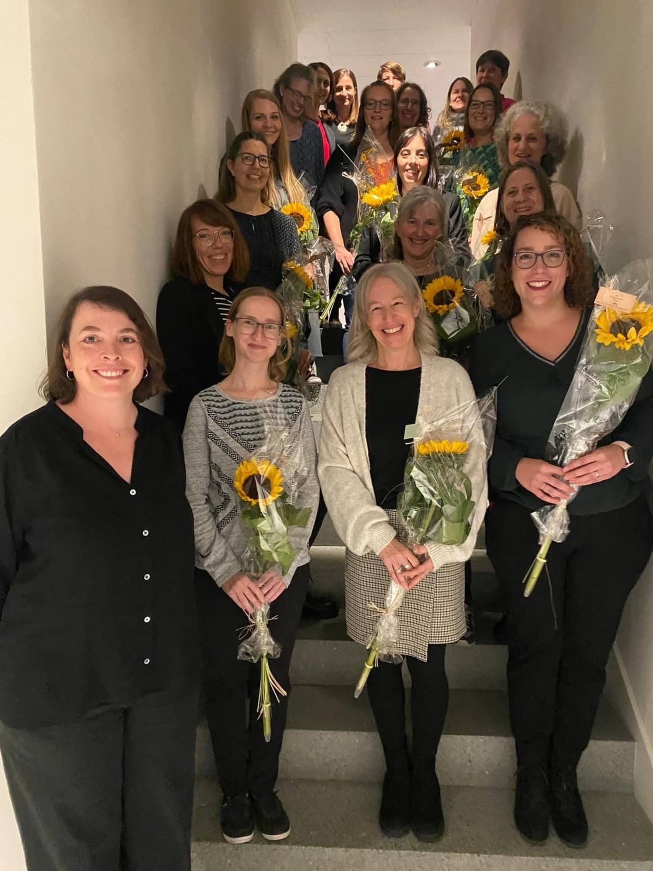 20 Frauen stehen für ein Gruppenfoto auf einer Treppe und halten Sonnenblumen in der Hand.