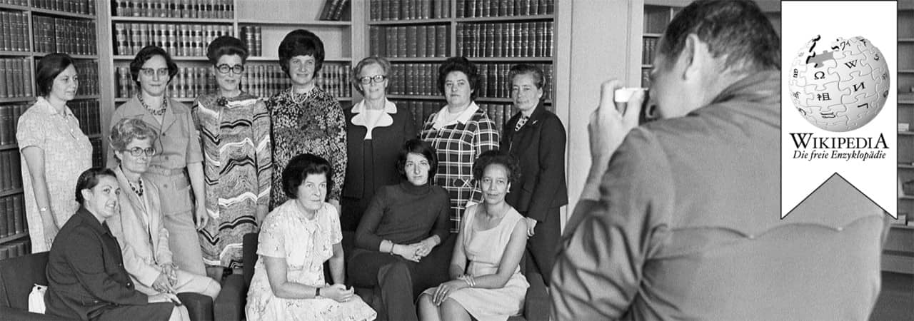 Gruppenfoto in Schwarzweiss mit zwölf Frauen sitzend und stehen. Auf dem Foto ist auch ein Fotograf von hinten zu sehen. Er fotografiert die Gruppe mit einer kleinen Kamera.