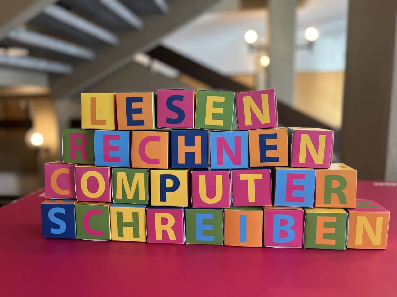 Bunte Buchstabenwürfel aus Papier liegen gestapelt auf einem roten Tisch und bilden die Worte Lesen, Rechnen, Computer und Schreiben.