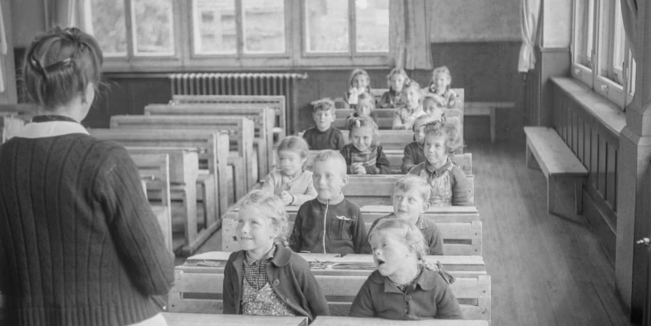 Schwarzweiss-Foto zeigt eine Schulklasse mit Blick zur Lehrerin, die mit dem Rücken zur Kamera steht