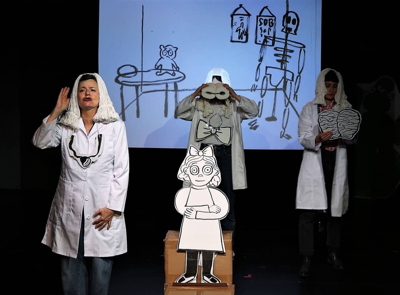 Drei Darstellerinnen mit weissem Kittel und weisser Perrücke umgeben ein Mädchen aus Papier. Im Hintergrund eine projizierte Kinderzeichnung.