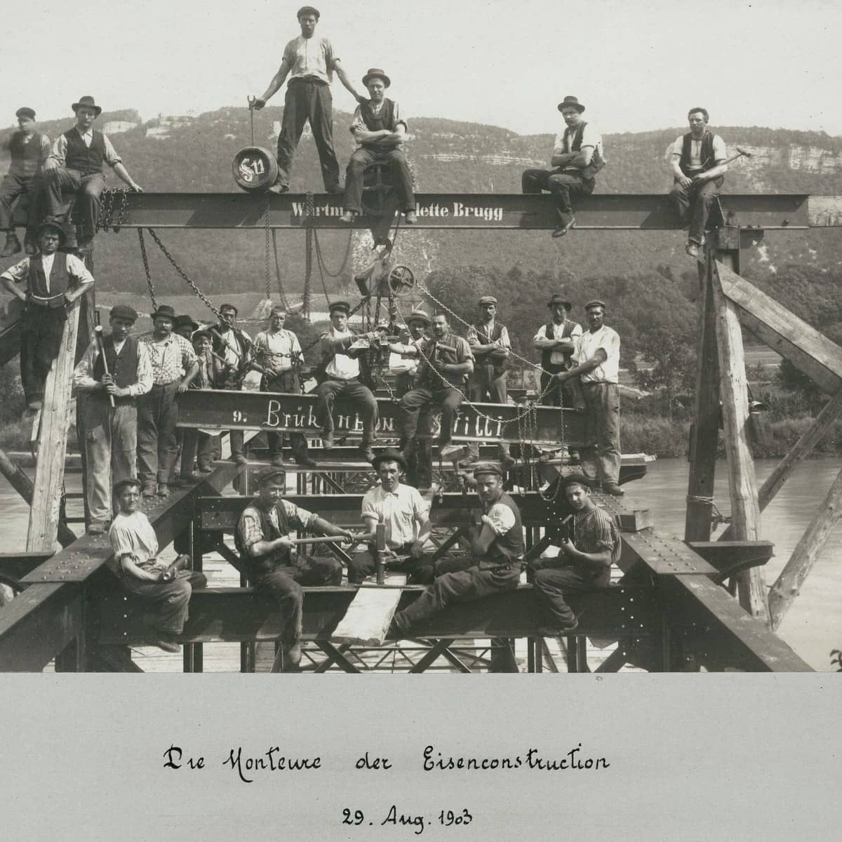 Schwarzweisses Foto zeigt Monteure beim Brückenbau auf einem Eisengerüst sitzend und stehend mit Werkzeugen in der Hand. Im Hintergrund die Aare und Hügelkette zu sehen. 