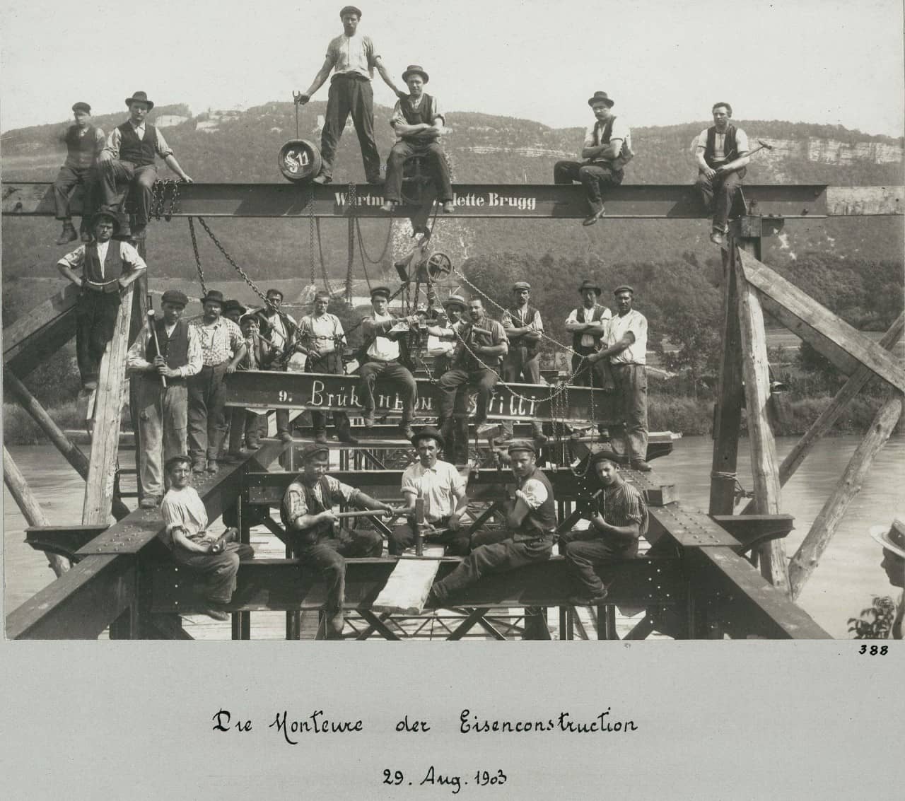 Schwarzweisses Foto zeigt Monteure beim Brückenbau auf einem Eisengerüst sitzend und stehend mit Werkzeugen in der Hand. Im Hintergrund die Aare und Hügelkette zu sehen. 