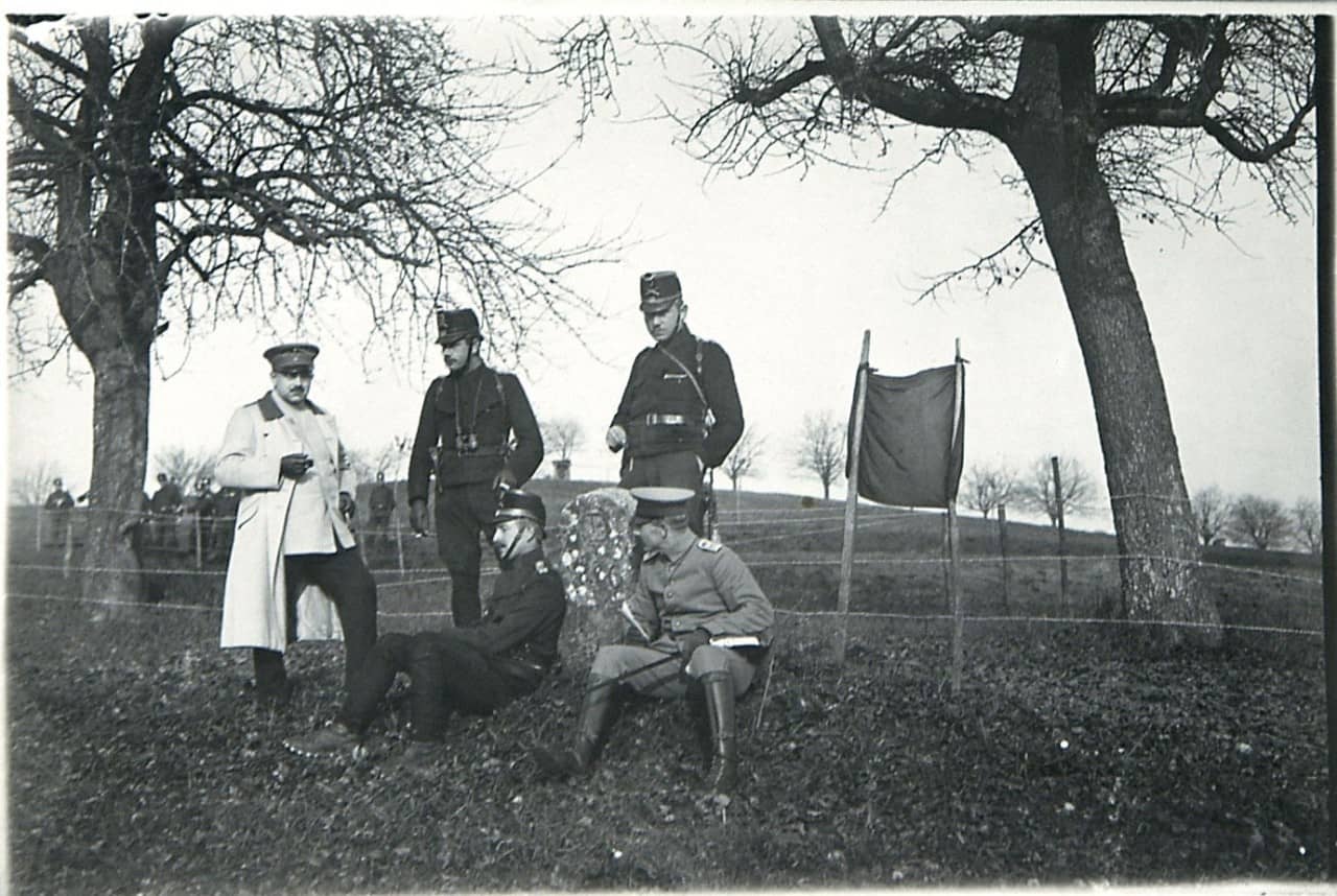 Schwarzweisses Foto zeigt fünf Offiziere, drei hinten stehend, zwei vorne sitzend an Grenzstein lehnend. Sie befinden sich draussen zwischen zwei Bäumen. Im Hintergrund eine Gruppe von Soldaten und weitere Bäume zu sehen. 