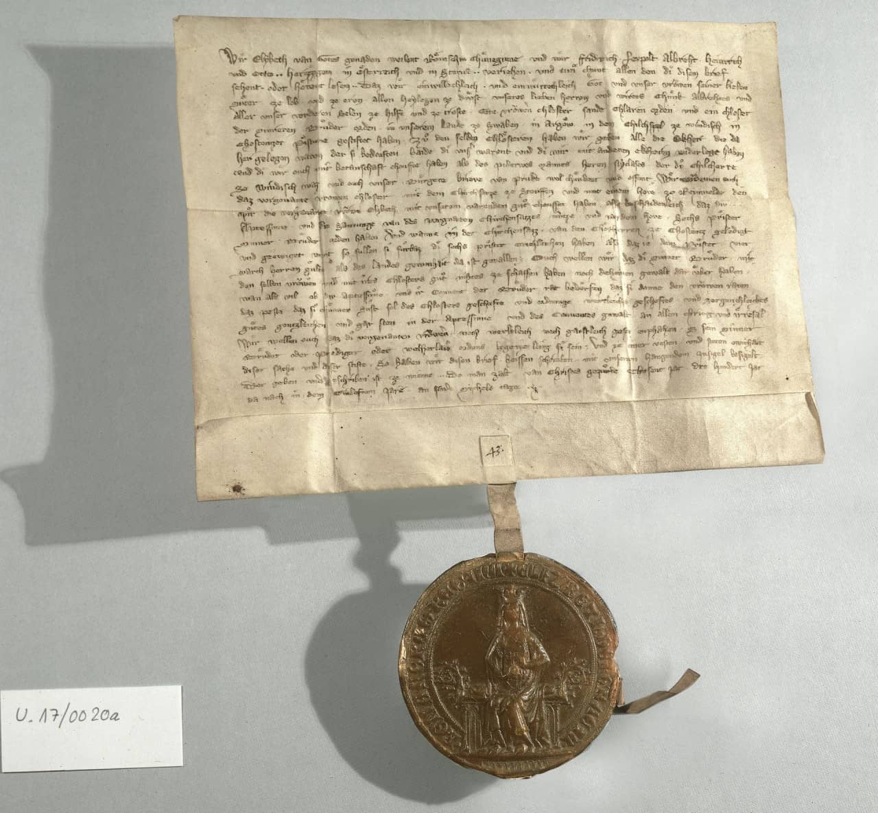 Pergament Urkunde mit dunkler Schrift und nach unten hängendem Siegel welches Königin auf Thron zeigt. Urkunde und Siegel werfen nach links einen Schatten.