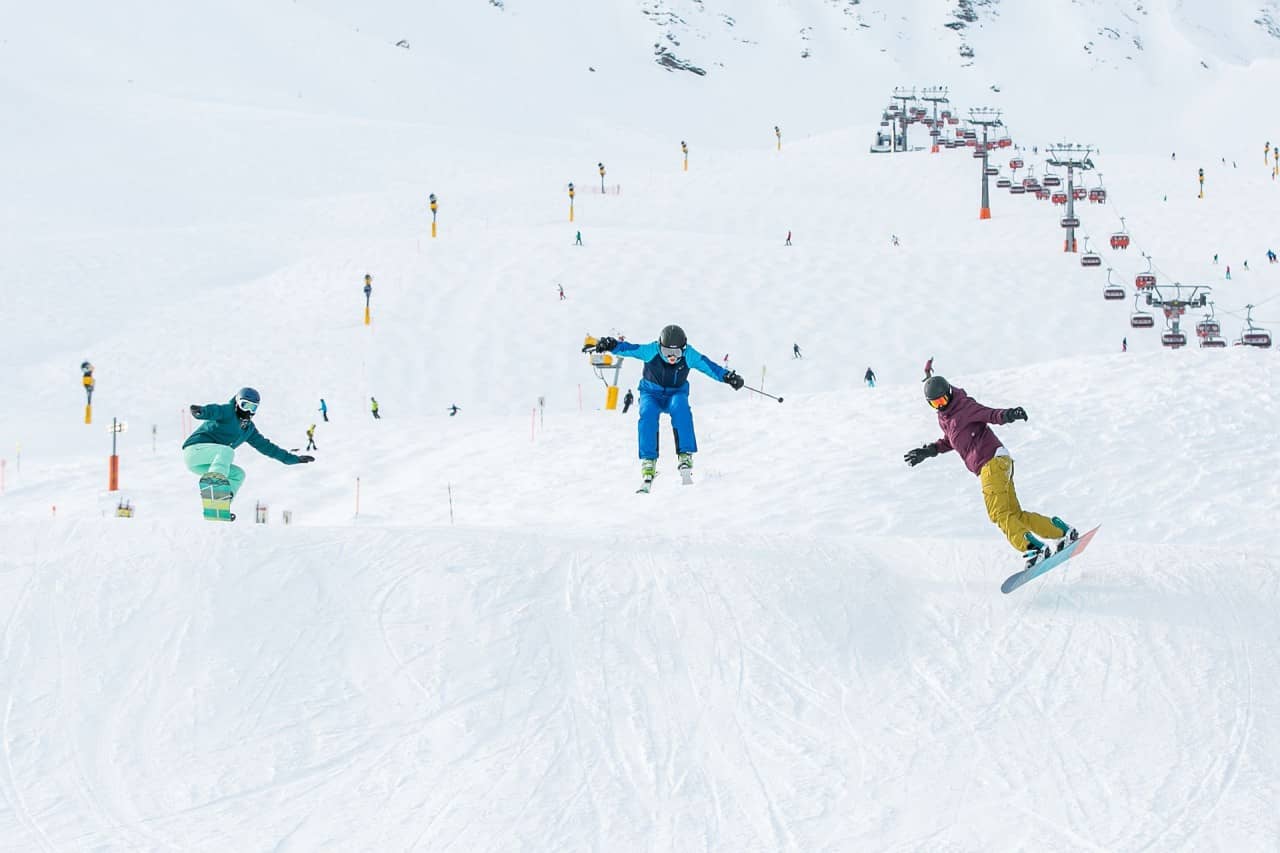 Ein Skifahrer/in und zwei Snowboarder/innen fahren auf der Piste den Hang hinunter.